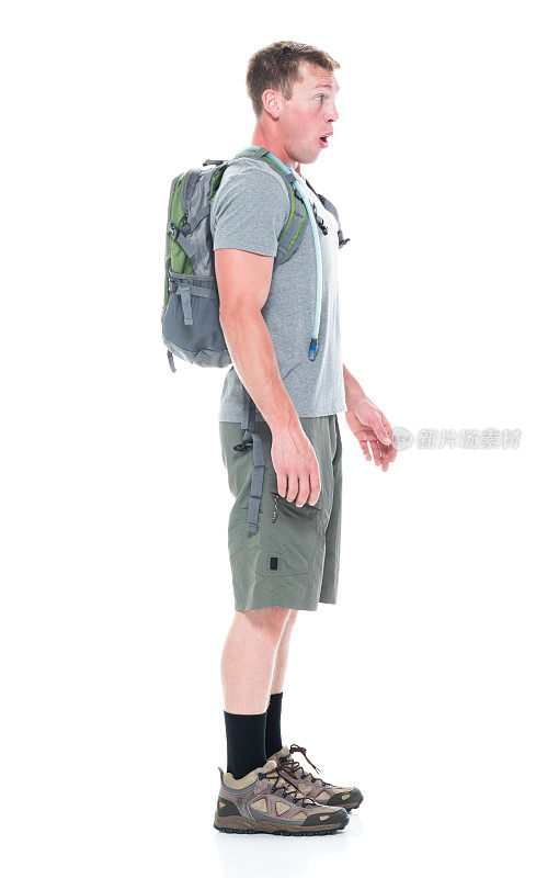 侧视图/全长20-29岁的白人青年男子/男性背包客徒步旅行/站在白色背景前，穿着短裤/背包/徒步靴/ t恤谁是在震惊/惊讶谁是探索和拿着包/探险家